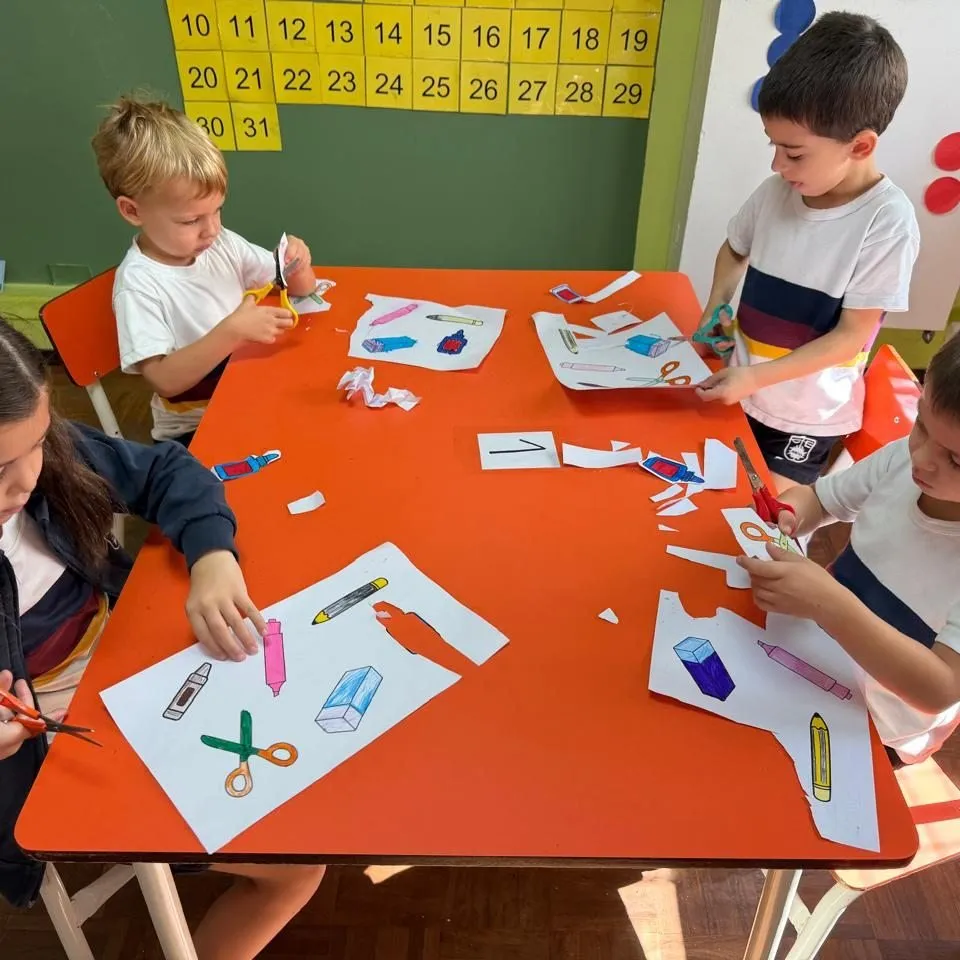 Manos de nenes de inicial recortando útiles de papel en una mesa naranja