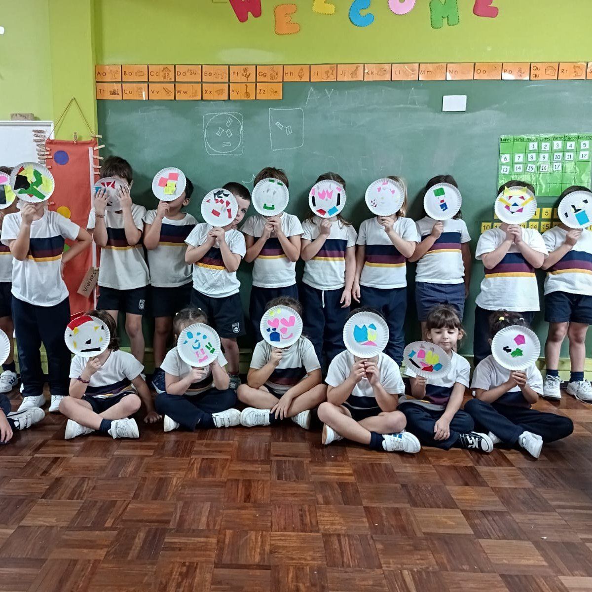 Alumnos con máscaras de platos de papel frente al pizarrón de Welcome, en clase de inglés
